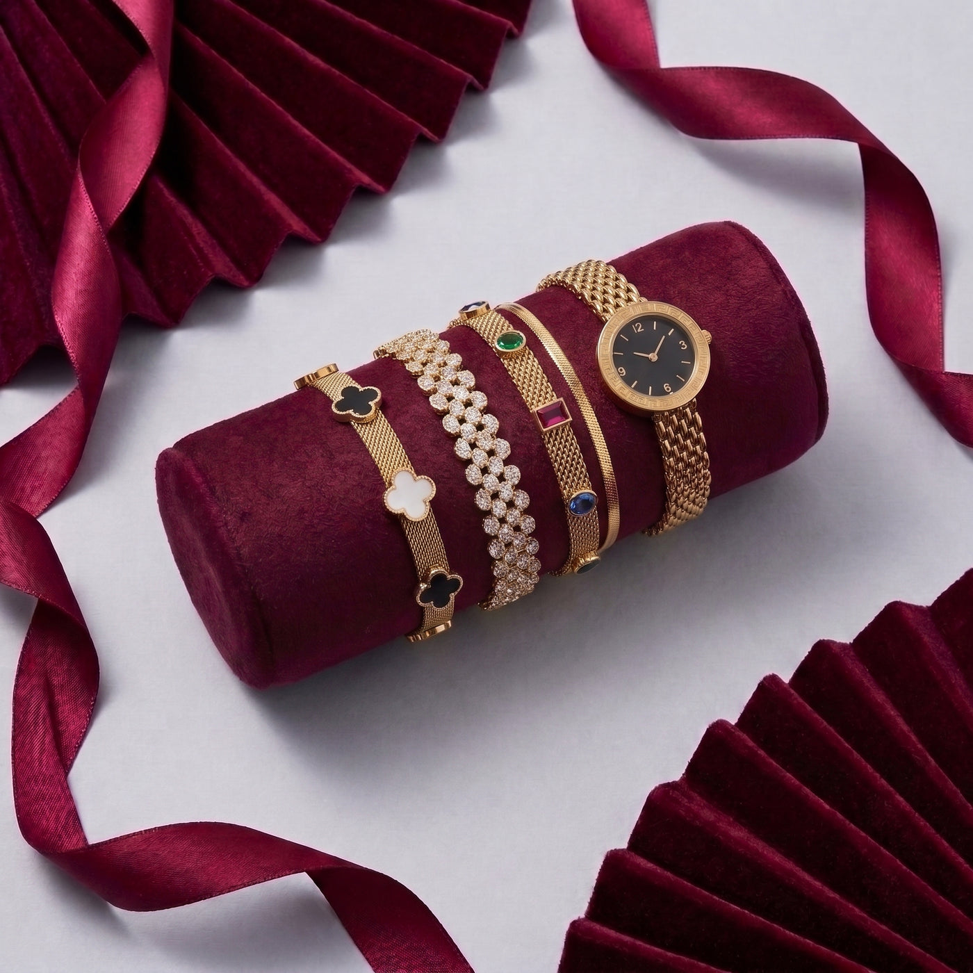 A stack of gold-toned bracelets and a matching watch displayed on a cylindrical burgundy velvet stand, surrounded by draped burgundy ribbon and folded matching fabric on a light gray background.