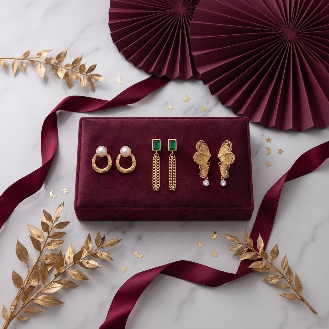 A set of four gold-toned earrings displayed on a burgundy velvet jewelry box, styled with gold decorative leaves, ribbons, and paper fans on a light marble surface.