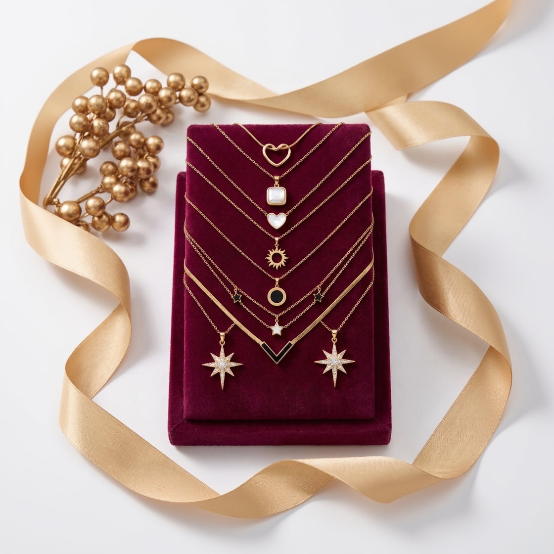 A set of gold-toned layered necklaces with various pendants (hearts, stars, sun, and geometric shapes) displayed on a deep burgundy velvet jewelry box. The box is styled with a gold ribbon and a decorative gold berry branch on a clean white background.