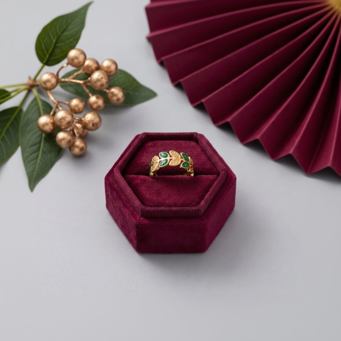 A gold ring with green and red enamel details sits in a hexagonal burgundy velvet ring box, styled with green leaves, gold berries, and a matching burgundy pleated fan on a light gray background.